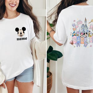 May include: White t-shirt featuring Mickey Mouse's face with the word "name" below it. The back of the shirt has a colorful graphic of a castle, characters, and the word "MAGICAL".