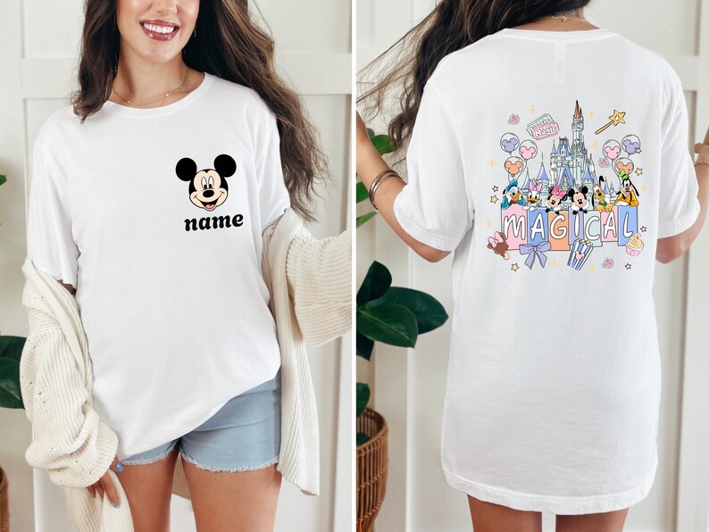 May include: White t-shirt featuring Mickey Mouse's face with the word "name" below it. The back of the shirt has a colorful graphic of a castle, characters, and the word "MAGICAL".