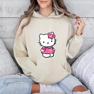 May include: A beige hooded sweatshirt featuring a central graphic of a white cartoon cat character wearing a bright pink glittery dress and a matching pink glittery bow on its head. The hoodie includes a front kangaroo pocket and long sleeves.
