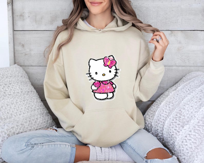 May include: A beige hooded sweatshirt featuring a central graphic of a white cartoon cat character wearing a bright pink glittery dress and a matching pink glittery bow on its head. The hoodie includes a front kangaroo pocket and long sleeves.