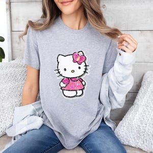 May include: A heather gray short-sleeved t-shirt featuring a central graphic of the character Hello Kitty. The character is depicted with a bright pink glittery dress and a matching pink glittery bow on its head, outlined in a dark border.