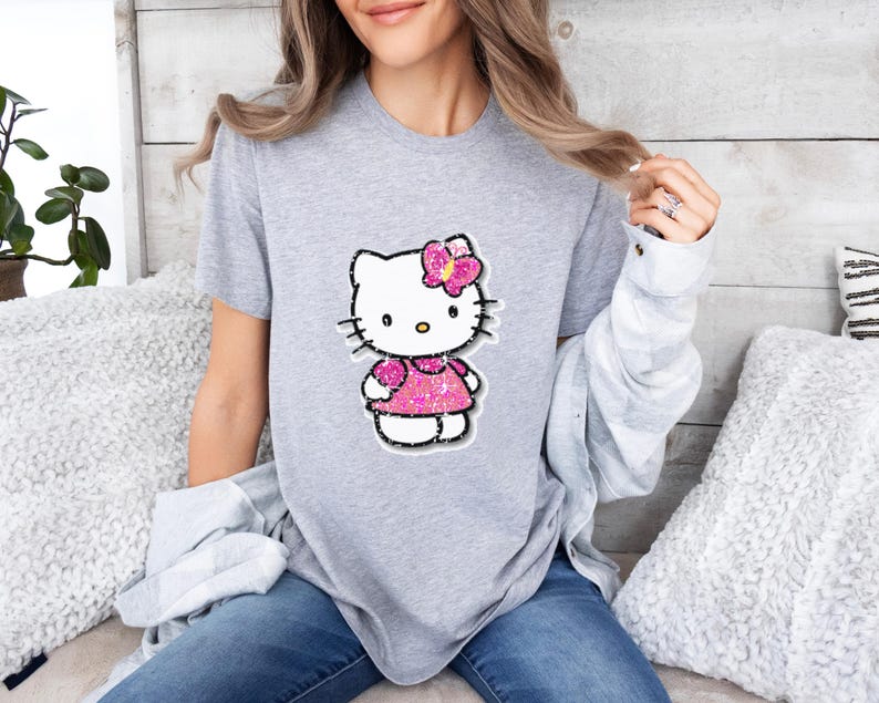 May include: A heather gray short-sleeved t-shirt featuring a central graphic of the character Hello Kitty. The character is depicted with a bright pink glittery dress and a matching pink glittery bow on its head, outlined in a dark border.