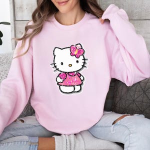 May include: A light pink crewneck sweatshirt featuring a central graphic of the character Hello Kitty. The character is depicted with a white face, black whiskers, and a bright pink glittery dress and matching bow on the side of the head.