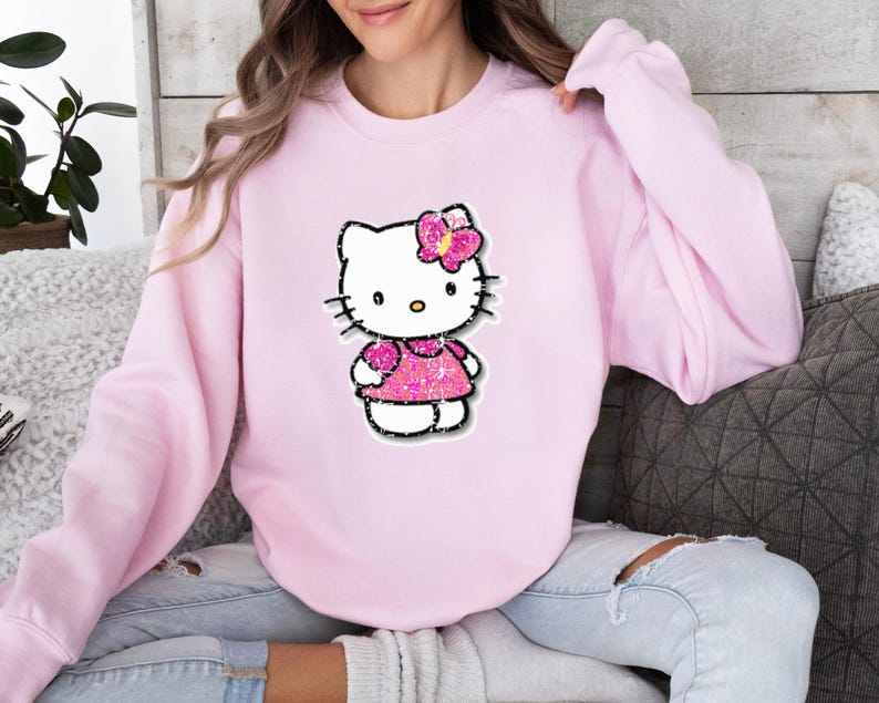 May include: A light pink crewneck sweatshirt featuring a central graphic of the character Hello Kitty. The character is depicted with a white face, black whiskers, and a bright pink glittery dress and matching bow on the side of the head.