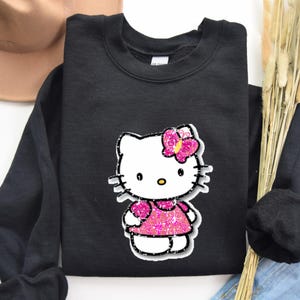 May include: A black long-sleeve sweatshirt featuring a graphic print of a white cat character with a yellow nose and black whiskers. The character wears a bright pink glittery dress and a matching pink glittery butterfly hair accessory.