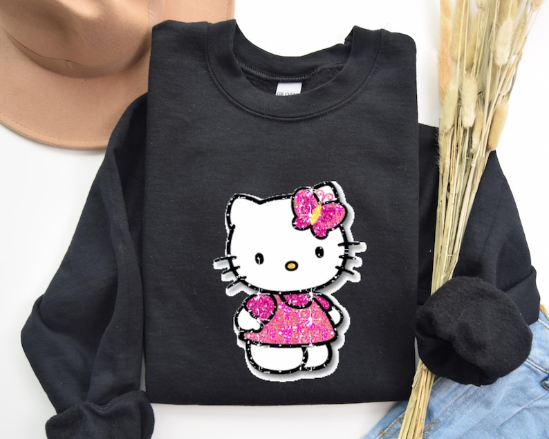 May include: A black long-sleeve sweatshirt featuring a graphic print of a white cat character with a yellow nose and black whiskers. The character wears a bright pink glittery dress and a matching pink glittery butterfly hair accessory.