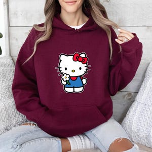 Pu&ograve; includere: Una felpa con cappuccio color bordeaux con una stampa grafica del personaggio Hello Kitty sul davanti. Il personaggio &egrave; bianco con un fiocco rosso, indossa un completo blu e tiene in mano un piccolo fiore bianco con il centro giallo.
