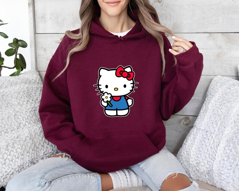 Pu&ograve; includere: Una felpa con cappuccio color bordeaux con una stampa grafica del personaggio Hello Kitty sul davanti. Il personaggio &egrave; bianco con un fiocco rosso, indossa un completo blu e tiene in mano un piccolo fiore bianco con il centro giallo.