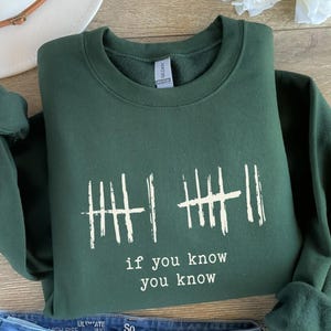 May include: A dark green sweatshirt with a crew neck. The front of the sweatshirt features a white graphic of tally marks and the text "if you know you know". The sweatshirt is folded.