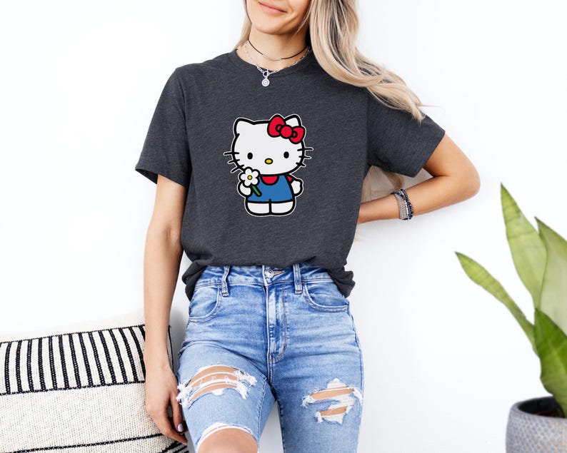 Pu&ograve; includere: Una t-shirt a maniche corte grigio antracite con una stampa grafica del personaggio Hello Kitty. Il personaggio &egrave; bianco con un fiocco rosso, indossa una salopette blu e tiene un piccolo fiore bianco con il centro giallo sul petto.