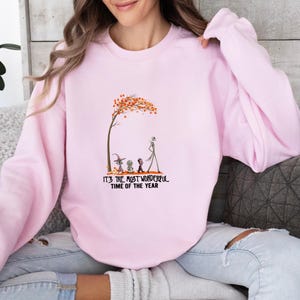 May include: A light pink sweatshirt with a graphic featuring characters under a tree with orange leaves. The text "IT'S THE MOST WONDERFUL TIME OF THE YEAR" is printed below the graphic. The sweatshirt is worn by a person.