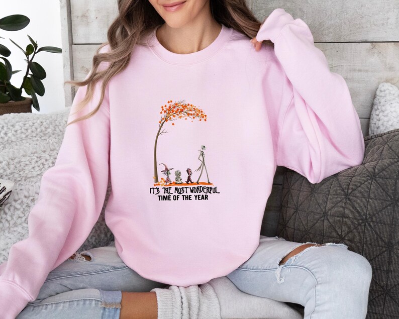 May include: A light pink sweatshirt with a graphic featuring characters under a tree with orange leaves. The text "IT'S THE MOST WONDERFUL TIME OF THE YEAR" is printed below the graphic. The sweatshirt is worn by a person.