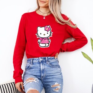 May include: A long-sleeve red crewneck sweatshirt featuring a central graphic of the character Hello Kitty. The character is depicted with a pink glittery dress and a matching pink glittery bow on its head, set against a white background.