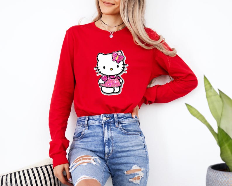 May include: A long-sleeve red crewneck sweatshirt featuring a central graphic of the character Hello Kitty. The character is depicted with a pink glittery dress and a matching pink glittery bow on its head, set against a white background.