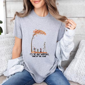May include: A heather gray t-shirt featuring a whimsical illustration of characters under a tree with orange leaves. The text "IT'S THE MOST WONDERFUL TIME OF THE YEAR" is printed below the image. The shirt is a casual, everyday wear item.