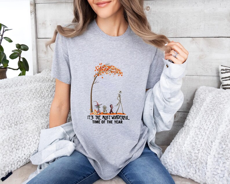 May include: A heather gray t-shirt featuring a whimsical illustration of characters under a tree with orange leaves. The text "IT'S THE MOST WONDERFUL TIME OF THE YEAR" is printed below the image. The shirt is a casual, everyday wear item.
