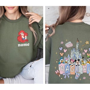 May include: Olive green sweatshirt featuring a cartoon image of Ariel with the word "name" below. The back of the sweatshirt has a colorful graphic of a castle, Disney characters, and the word "MAGICAL".