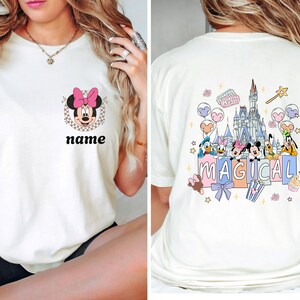 May include: Cream-colored t-shirt featuring Minnie Mouse with a pink bow and the text "name" on the front. The back of the shirt showcases a colorful graphic of Disney characters and a castle with the word "MAGICAL".