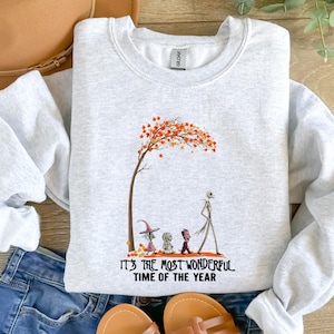 May include: Light gray sweatshirt with a graphic of a tree with orange leaves and cartoon characters. The text "IT'S THE MOST WONDERFUL TIME OF THE YEAR" is printed below the graphic. The sweatshirt is paired with blue jeans and sandals.