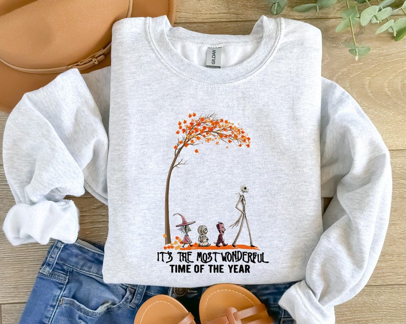 May include: Light gray sweatshirt with a graphic of a tree with orange leaves and cartoon characters. The text "IT'S THE MOST WONDERFUL TIME OF THE YEAR" is printed below the graphic. The sweatshirt is paired with blue jeans and sandals.