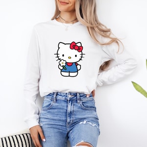Pu&ograve; includere: Una maglietta bianca a maniche lunghe con la grafica di un personaggio gatto bianco con un fiocco rosso, che indossa un completo blu e tiene in mano un piccolo fiore bianco con il centro giallo. La maglia ha un girocollo e polsini a coste.