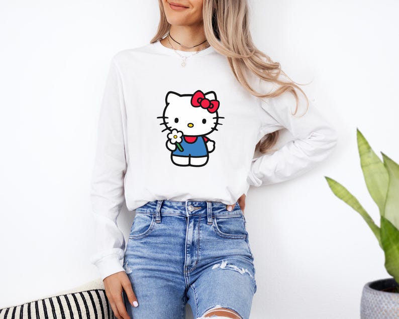 Pu&ograve; includere: Una maglietta bianca a maniche lunghe con la grafica di un personaggio gatto bianco con un fiocco rosso, che indossa un completo blu e tiene in mano un piccolo fiore bianco con il centro giallo. La maglia ha un girocollo e polsini a coste.