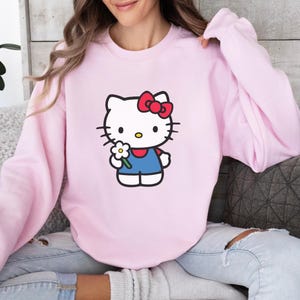 Pu&ograve; includere: Una felpa girocollo rosa chiaro con una stampa grafica di Hello Kitty. Il personaggio &egrave; bianco con un fiocco rosso, indossa un completo blu e tiene in mano un piccolo fiore bianco con il centro giallo. La felpa ha maniche lunghe e collo a coste.