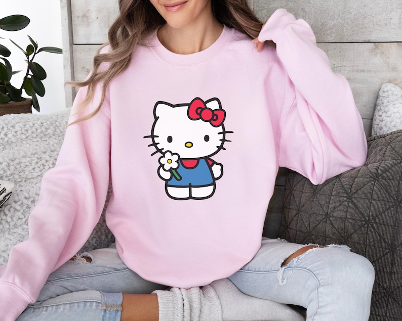 Pu&ograve; includere: Una felpa girocollo rosa chiaro con una stampa grafica di Hello Kitty. Il personaggio &egrave; bianco con un fiocco rosso, indossa un completo blu e tiene in mano un piccolo fiore bianco con il centro giallo. La felpa ha maniche lunghe e collo a coste.