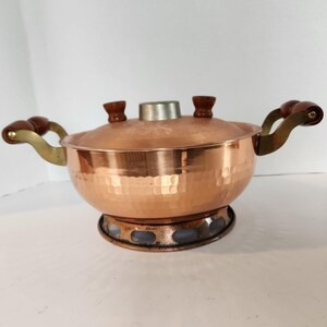 May include: A copper pot with a lid and two handles. The pot has a hammered texture and a round base. The handles are made of wood and brass. The lid has two small wooden knobs and a silver metal center.