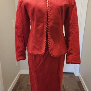 Puede incluir: Un traje de falda rojo con ribete festoneado. La chaqueta tiene un escote sin cuello y mangas largas. La falda es de largo hasta la rodilla.