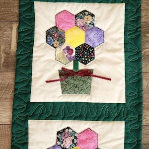 May include: A quilted table runner featuring three floral arrangements. Each arrangement has a green pot with a burgundy bow, a green stem, and a collection of colorful, hexagonal flower petals. The border is green with a decorative stitch.
