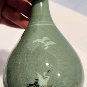 May include: A celadon ceramic vase with a crackle glaze. The vase features a painted crane design and cloud motifs. The vase is held by a hand.