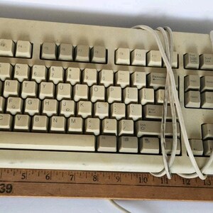 May include: A beige Dell QuietKey computer keyboard with a QWERTY layout and a coiled cable. A wooden ruler is positioned along the bottom edge, showing the keyboard measures approximately 18 inches in total length from end to end.