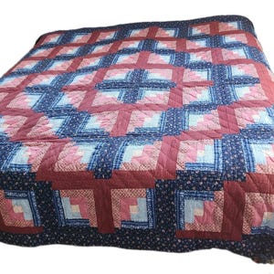 May include: A handmade quilt with a patchwork design. The quilt features a pattern of squares in shades of red, blue, and pink. The border is a dark blue floral print. The quilt is likely made of cotton and is suitable for a bed.