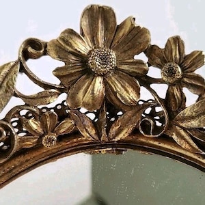 May include: Ornate gold-toned mirror with a floral design. The top of the mirror features detailed flowers and leaves. The mirror's frame has an oval shape and a decorative edge.