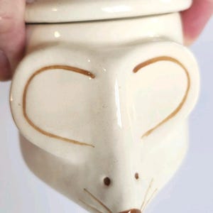 May include: A cream-colored ceramic jar with a lid, shaped like a mouse head. The jar features large, rounded ears outlined in brown, two small brown dots for eyes, and a brown nose with thin brown whiskers painted on the front surface.