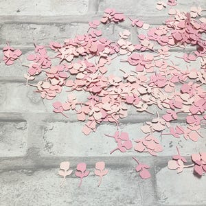 May include: A large pile of pink paper flower confetti. The confetti is made of paper and is in the shape of small flowers with stems. The confetti is scattered on a gray brick background.