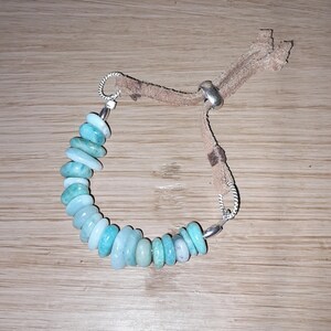 Pode incluir: Um bracelete de pedras turquesa com um cordão de couro castanho e fechos de prata. O bracelete é feito de pedras redondas e planas.