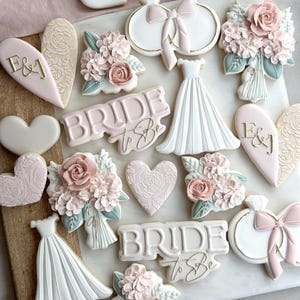 May include: A collection of decorated sugar cookies featuring a bridal theme. The cookies are decorated with pink, white, and gold icing and feature designs such as hearts, flowers, a wedding dress, and the words "Bride to Be".