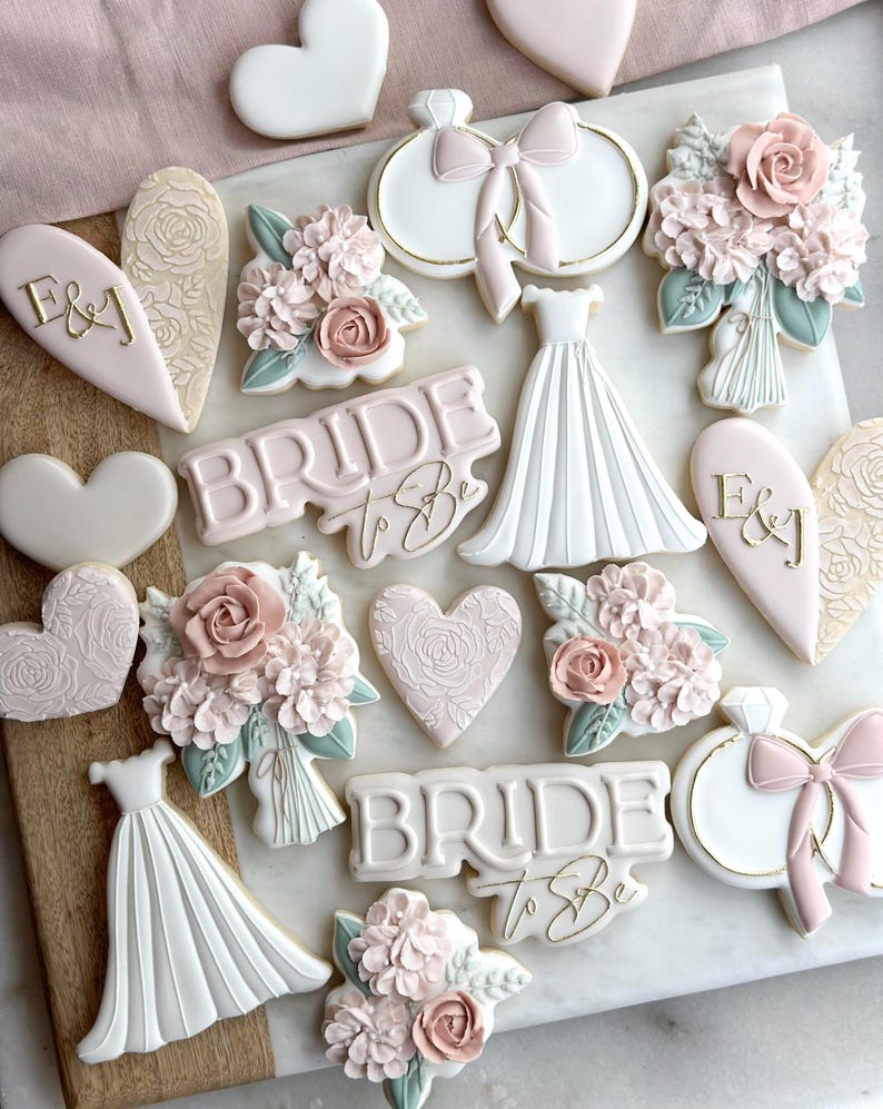 May include: A collection of decorated sugar cookies featuring a bridal theme. The cookies are decorated with pink, white, and gold icing and feature designs such as hearts, flowers, a wedding dress, and the words "Bride to Be".