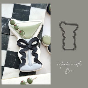 May include: A cookie cutter in the shape of a martini glass with a bow. The cutter is made of metal and is gray in color. The text "Martini with Bow" is written below the cutter.