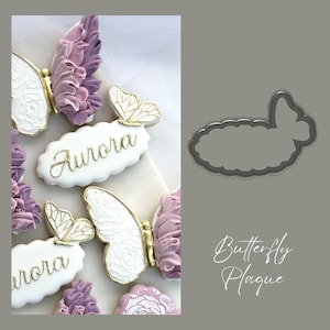 Può includere: Un primo piano di uno stampino per biscotti a forma di farfalla. Lo stampino è in metallo e ha una finitura argentata. Lo stampino è su uno sfondo bianco. Il testo "Butterfly Plaque" è scritto in corsivo nero sotto lo stampino.