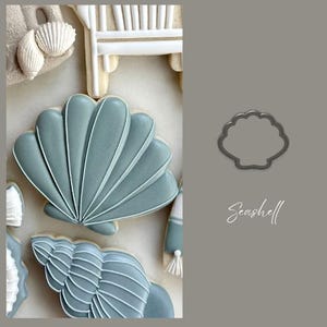 May include: Cookies decorated in a beach theme. The cookies are in the shape of seashells, a beach chair, and a seashell cookie cutter. The cookies are light blue and white.