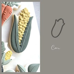 May include: A decorated cookie shaped like an ear of corn, featuring a light yellow kernel design and green husks. The cookie is part of a set. The word "Corn" is written in cursive.