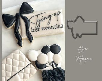 May include: A set of three cookie cutters, one shaped like a bow, one shaped like a plaque, and one shaped like a present with a bow. The present cookie cutter has the text "Tying up her twenties" on it.