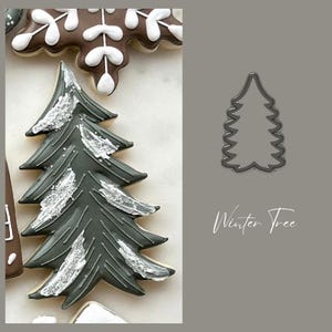 May include: A green and white frosted sugar cookie shaped like a Christmas tree. The cookie is decorated with white icing that looks like snow. The cookie is next to a metal cookie cutter shaped like a Christmas tree. The text "Winter Tree" is written below the cookie cutter.