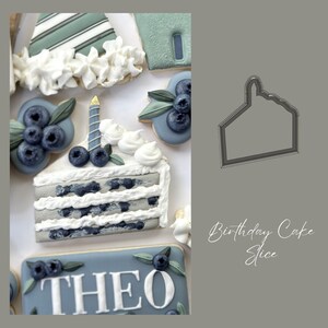 May include: A blue and white decorated sugar cookie shaped like a slice of cake with blueberries and a candle on top. The cookie is decorated with a gray and white icing and has the text "Birthday Cake Slice" written below it.