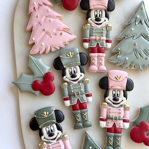 May include: A collection of decorated sugar cookies featuring Mickey Mouse dressed as a nutcracker in pink, green, and red icing, alongside pink and green Christmas tree cookies and holly leaves with red Mickey Mouse head accents.