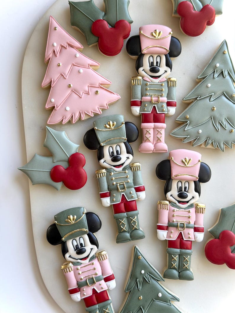 May include: A collection of decorated sugar cookies featuring Mickey Mouse dressed as a nutcracker in pink, green, and red icing, alongside pink and green Christmas tree cookies and holly leaves with red Mickey Mouse head accents.