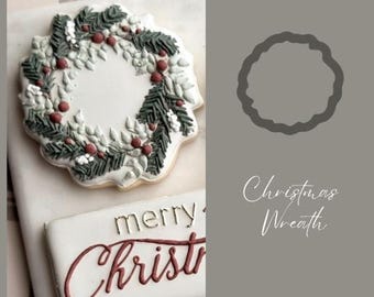 May include: A decorated gingerbread cookie in the shape of a Christmas wreath with green leaves, red berries, and white accents. The cookie is on a white surface with a smaller cookie below it that says "merry Christmas" in red and gold lettering.
